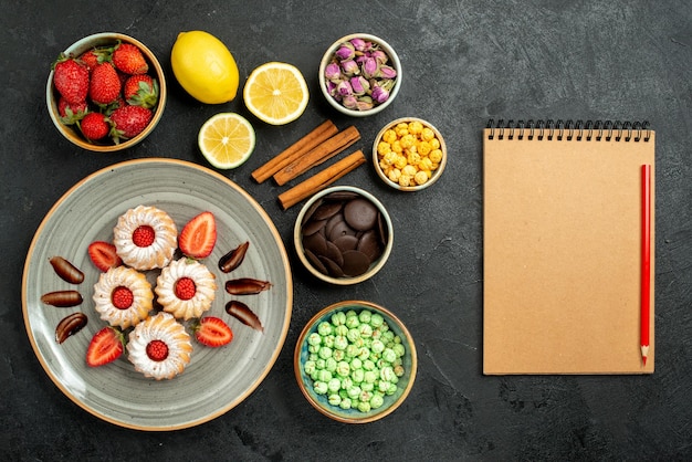 Vista dall'alto da lontano dolci con biscotti da tè con tè nero alla fragola con nocciole al limone ciotole di cioccolato e dolci diversi accanto a quaderno crema e matita rossa su superficie scura