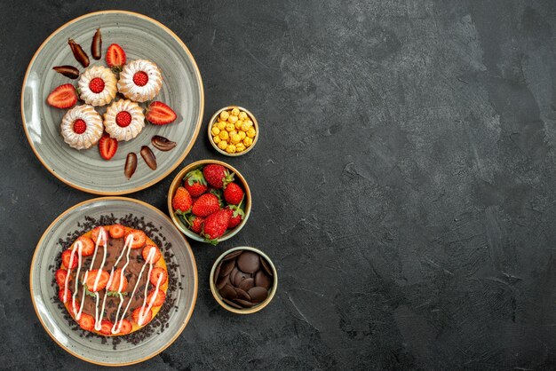 Vista dall'alto da lontano dolci e torta appetitosi biscotti e torta con fragole e cioccolato e ciotole di nocciole cioccolato e fragola sul lato sinistro del tavolo nero