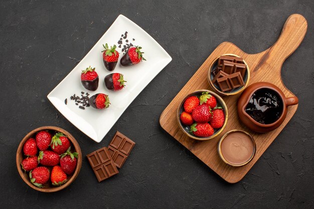 Vista dall'alto da lontano fragole ciotola di fragole barrette di cioccolato piatto di fragole ricoperte di cioccolato accanto alle ciotole di crema al cioccolato e fragole sul tagliere