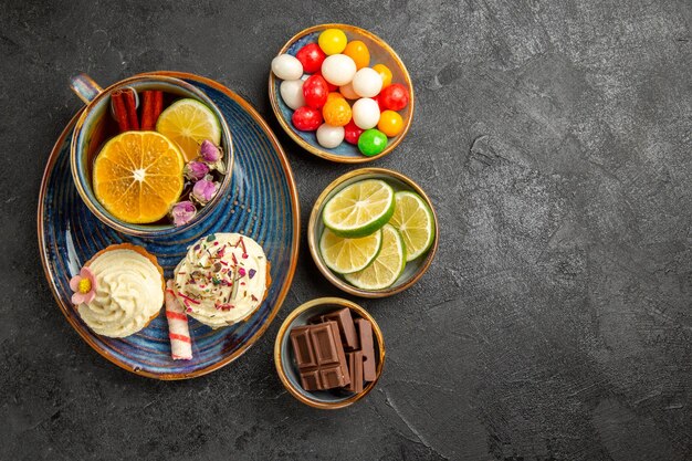 Vista dall'alto da lontano i dolci sul tavolo tre ciotole di caramelle al cioccolato e fette di lime accanto al piattino blu della tazza di tisana nera e due cupcakes sul tavolo