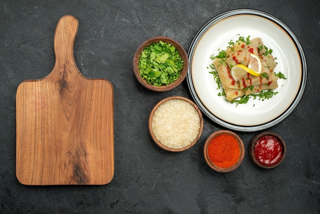 Vista dall'alto da lontano piatto con salsa di cavolo ripieno con erbe al limone e salsa su piatto bianco e spezie erbe di riso e salsa in ciotole accanto alla tavola da cucina in legno sul tavolo scuro