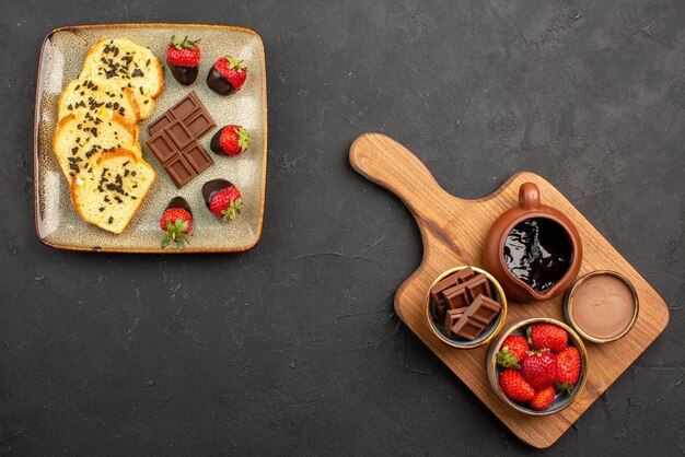 Vista dall'alto da lontano piatto da dessert di appetitosa torta con fragole ricoperte di cioccolato accanto a ciotole di crema al cioccolato e frutti di bosco sul tagliere sul tavolo scuro