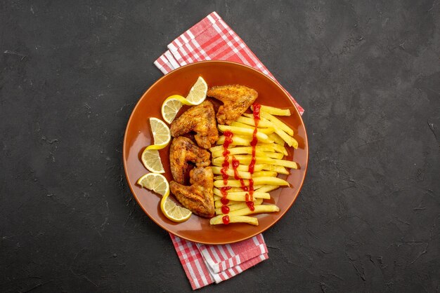 Vista dall'alto da lontano piatto su tovaglia piatto arancione di ali di pollo ketchup patatine fritte e pezzi di limone su tovaglia a quadretti rosa-bianco al centro del tavolo scuro