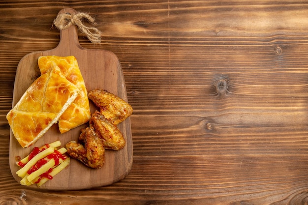 Vista dall'alto da lontano pollo e torta due pezzi di ali di pollo torta e patatine fritte con ketchup sul tagliere sul lato sinistro del tavolo scuro