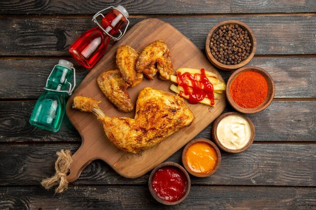 Vista dall'alto da lontano pollo rosso e blu bottiglie accanto al pollo con patatine fritte e ketchup sul tagliere ciotole di pepe nero salse spezie