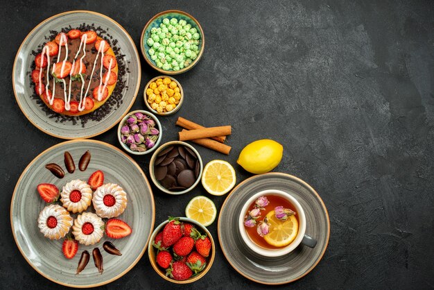 Vista dall'alto da lontano torta con dolci torta con tè nero alla fragola con limone piatto di biscotti con nocciole alla fragola ciotole di cioccolato e dolci diversi sul lato sinistro del tavolo scuro