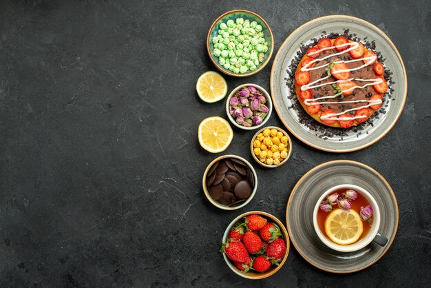 Vista dall'alto da lontano torta con tè appetitoso torta con cioccolato e tè nero alla fragola limoni ciotole di cioccolato e dolci diversi sul lato destro del tavolo nero