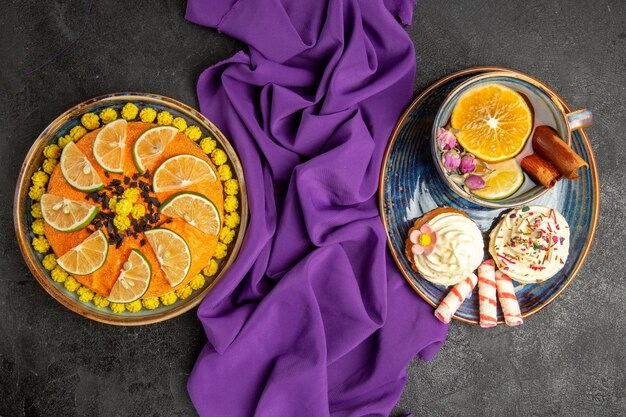 Vista dall'alto da lontano torta con torta appetitosa al limone con fette di arancia blu piatto di cupcakes con crema e una tazza di tè con bastoncini di cannella accanto alla tovaglia viola