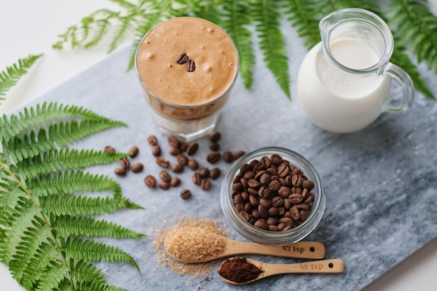 Vista dall'alto del caffè dalgona con latte di mandorla e zucchero di canna in grani di caffè e pianta verde su tavola di marmo Un bicchiere di soffice bevanda coreana alla moda con panna montata xD