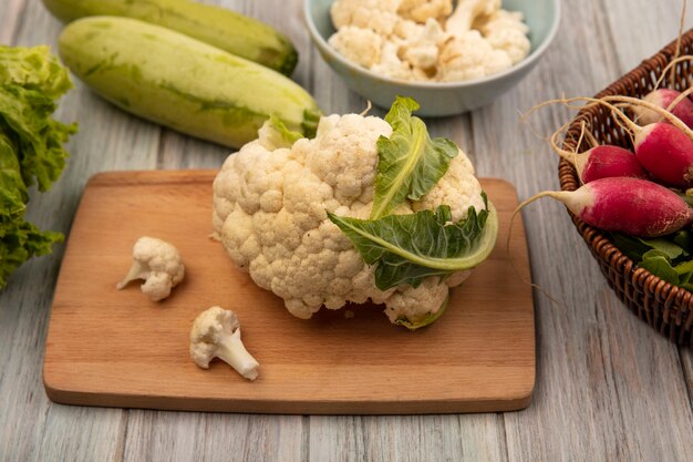 Vista dall'alto del cavolfiore ricco di vitamine su una tavola da cucina in legno con boccioli di cavolfiore su una ciotola con ravanelli su un secchio con zucchine isolato su una superficie di legno grigia