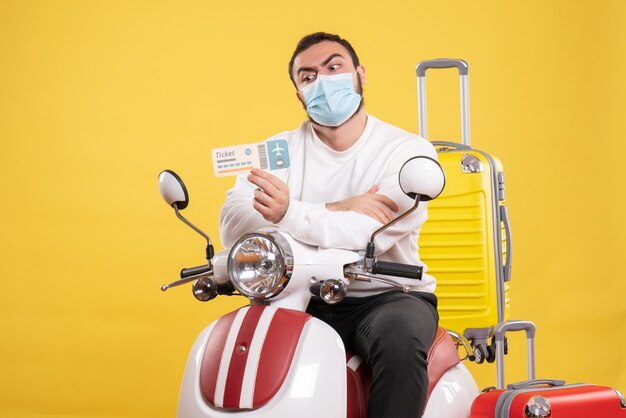 Vista dall'alto del concetto di viaggio con un giovane ragazzo in maschera medica seduto su una moto con una valigia gialla su di esso e con in mano il biglietto sentendosi scioccato