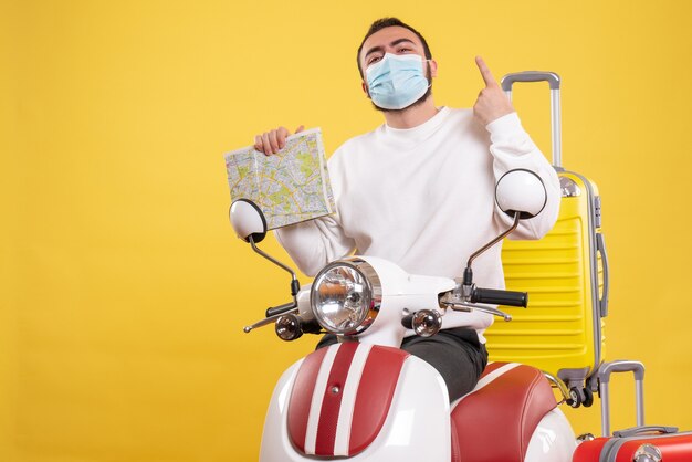 Vista dall'alto del concetto di viaggio con un ragazzo fiducioso in maschera medica in piedi vicino alla moto con valigia gialla su di essa e tenendo la mappa