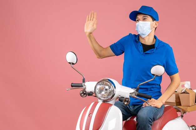 Vista dall'alto del corriere in maschera medica che indossa un cappello seduto su uno scooter che saluta qualcuno su sfondo color pesca pastello