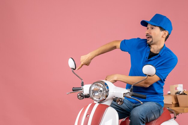 Vista dall'alto del corriere sorpreso che indossa un cappello seduto su uno scooter che consegna gli ordini che puntano verso il basso su una pesca pastello