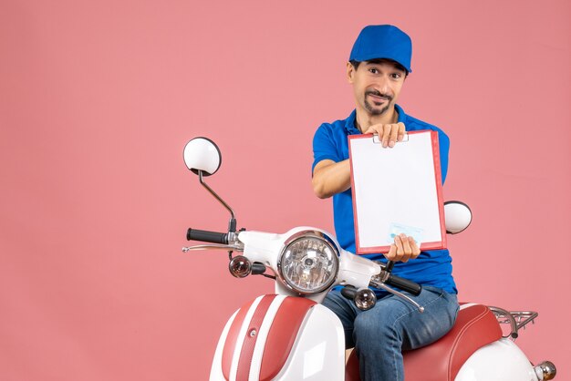 Vista dall'alto del corriere sorridente in maschera medica che indossa un cappello seduto su uno scooter che tiene un documento su una pesca pastello