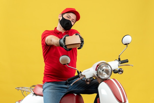 Vista dall'alto del corriere uomo che indossa camicia rossa e guanti cappello in mascherina medica che si siede sul motorino che dà ordine