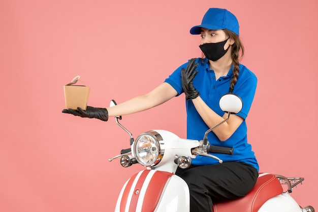 Vista dall'alto del fattorino confuso che indossa maschera medica e guanti seduto su uno scooter che consegna ordini su sfondo color pesca pastello