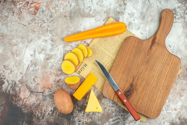 Vista dall'alto del fondo di cottura con varie verdure e due tipi di coltello da formaggio e tagliere di legno su un vecchio giornale su sfondo a colori misti