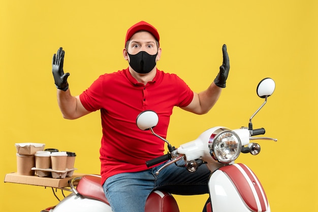 Vista dall'alto del giovane adulto che indossa camicetta rossa e guanti cappello in maschera medica offrendo ordine seduto sullo scooter sensazione scioccata su sfondo giallo