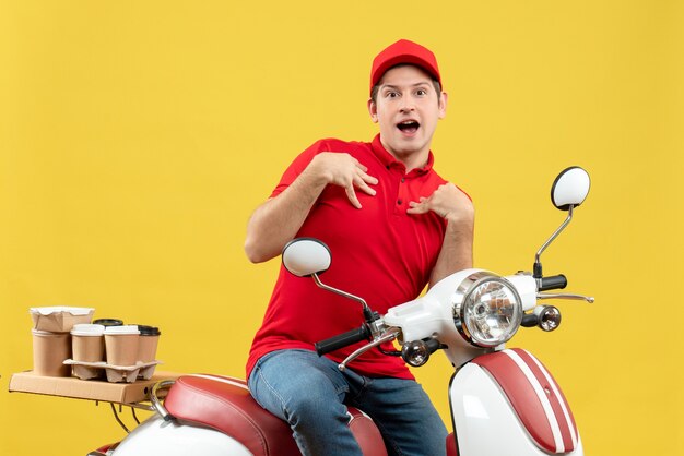 Vista dall'alto del giovane adulto confuso che indossa camicetta rossa e cappello offrendo ordine seduto su uno scooter che punta se stesso su sfondo giallo