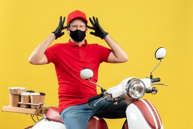 Vista dall'alto del giovane adulto emotivo che indossa guanti di camicetta e cappello rossi nella mascherina medica che trasporta l'ordine che si siede sullo scooter che soffre di mal di testa