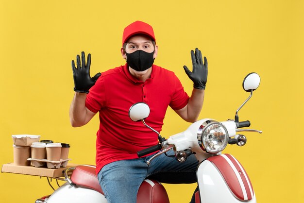 Vista dall'alto del giovane adulto laborioso che indossa guanti di camicetta e cappello rossi in maschera medica offrendo ordine seduto su scooter su sfondo giallo