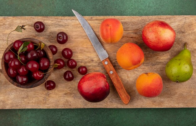 Vista dall'alto del modello di frutta come albicocche pesche pera ciliegie con ciotola di ciliegia e coltello sul tagliere su sfondo verde