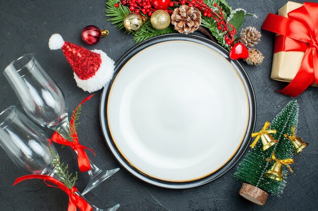 Vista dall'alto del piatto di cena albero di natale rami di abete conifera cono confezione regalo cappello di babbo natale calici di vetro caduti su sfondo nero