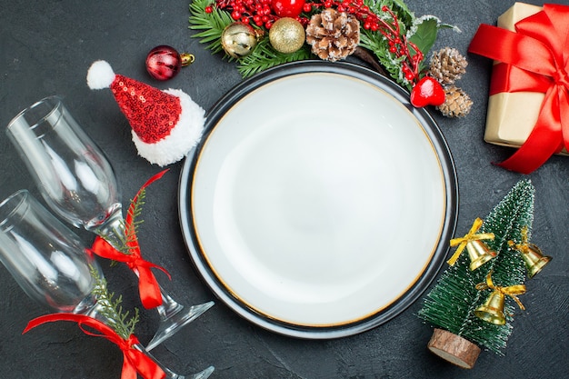 Vista dall'alto del piatto di cena albero di natale rami di abete conifera cono confezione regalo cappello di babbo natale calici di vetro caduti su sfondo nero