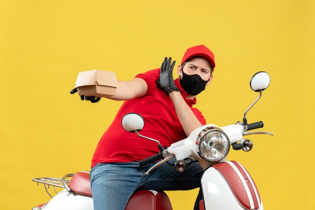 Vista dall'alto del ragazzo delle consegne impaurito che indossa guanti uniformi e cappello in maschera medica che si siede sullo scooter che mostra l'ordine