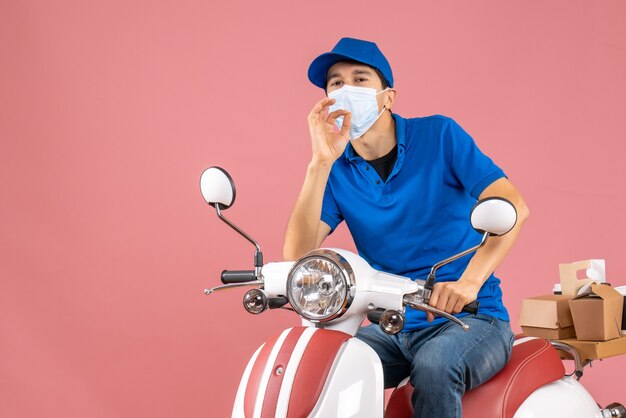 Vista dall'alto del ragazzo delle consegne in maschera medica che indossa un cappello seduto su uno scooter e fa un gesto perfetto su sfondo color pesca pastello