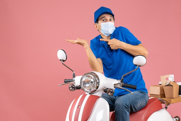 Vista dall'alto del ragazzo delle consegne sorpreso in maschera medica che indossa un cappello seduto su uno scooter su una pesca pastello