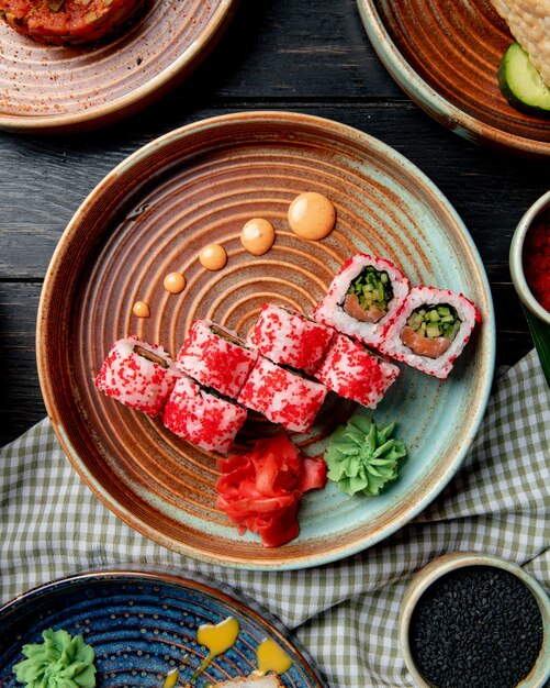 Vista dall'alto del rotolo di sushi con salmone avocado cetriolo e crema di formaggio ricoperto di caviale rosso con zenzero e wasabi su un piatto sul tavolo di legno