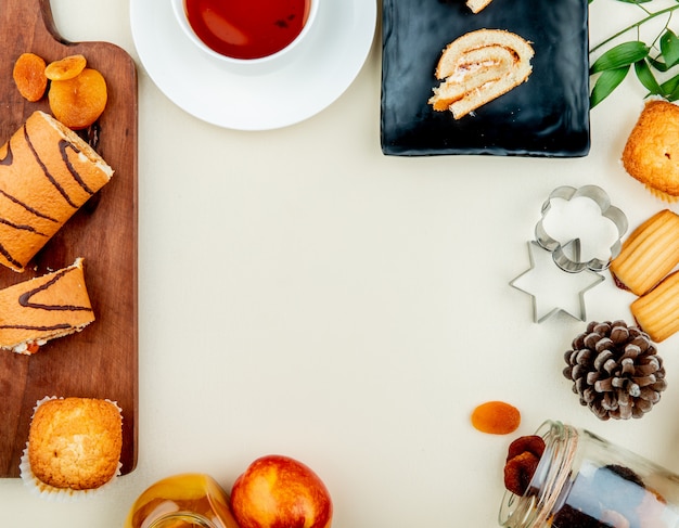 Vista dall'alto del rotolo tagliato e tranciato con prugne secche cupcake sul tagliere con marmellata di tè pesca uvetta biscotti e pigne su superficie bianca con spazio di copia