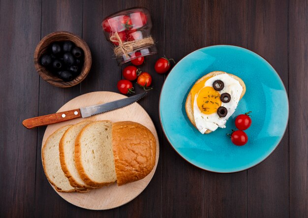 Vista dall'alto del set da colazione con fette di pane e coltello sul tagliere e piatto di uovo fritto con pomodori che fuoriescono dalla ciotola e ciotola di oliva nera su legno