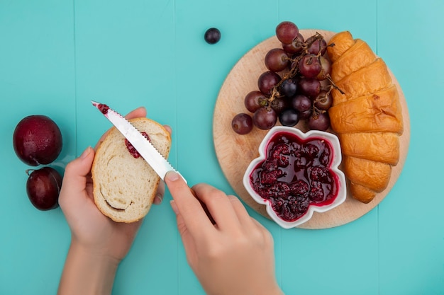Vista dall'alto del set per la colazione con croissant e marmellata di lamponi uva sul tagliere e mano femminile spalmando marmellata su pane e pluots su sfondo blu
