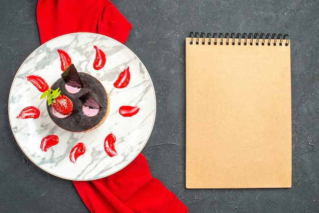 Vista dall'alto deliziosa cheesecake con fragole e cioccolato su piatto scialle rosso un quaderno su sfondo scuro isolato