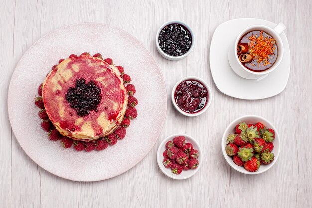 Vista dall'alto deliziose frittelle con fragole tazza di tè su torta bianca da scrivania biscotto dolce torta di frutti di bosco