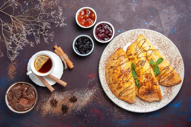 Vista dall'alto deliziosi pasticcini al forno con una tazza di marmellata di tè su uno spazio buio