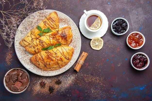 Vista dall'alto deliziosi pasticcini al forno con una tazza di marmellata di tè su uno spazio buio