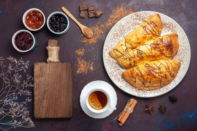 Vista dall'alto deliziosi pasticcini al forno con una tazza di tè e marmellata sulla scrivania scura