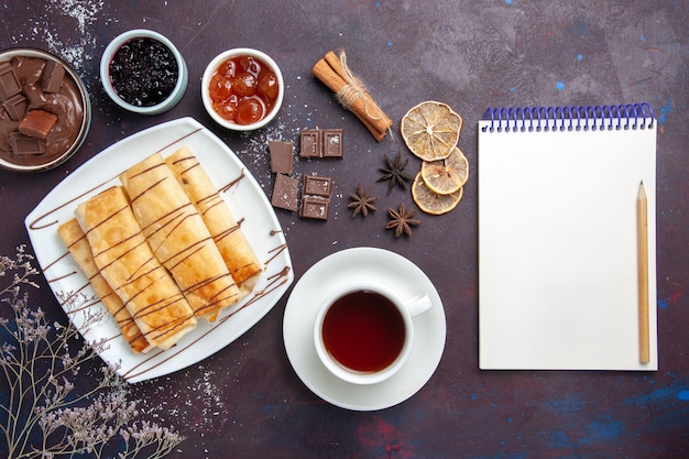 Vista dall'alto deliziosi pasticcini dolci con marmellata di cioccolato e tazza di tè sulla scrivania scura