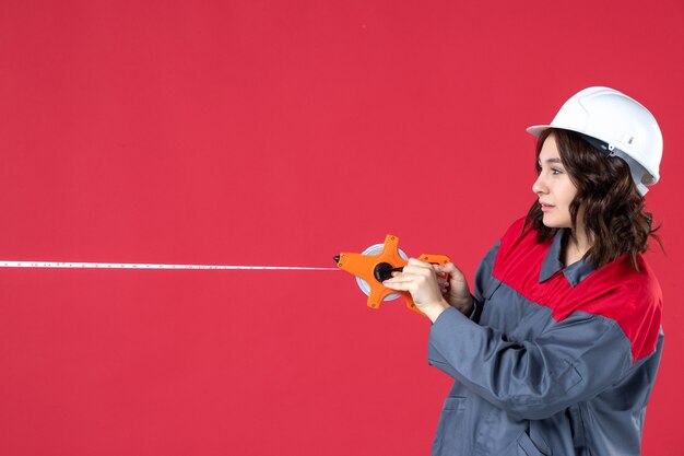 Vista dall'alto dell'architetto femminile sorridente occupato in uniforme con il nastro di misurazione dell'apertura del casco su sfondo rosso isolato