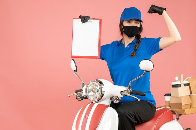 Vista dall'alto dell'orgogliosa ragazza del corriere che indossa maschera medica e guanti seduti su uno scooter con in mano un foglio di carta vuoto che consegna ordini su sfondo color pesca pastello