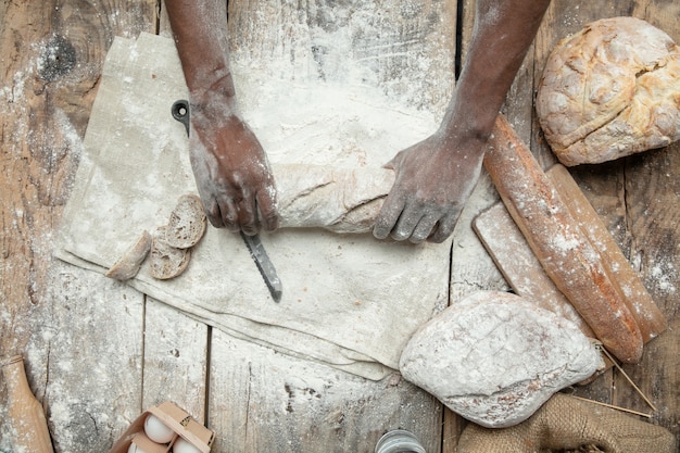 Vista dall'alto dell'uomo afro-americano cuochi cereali freschi, pane, crusca sulla tavola di legno. Mangiare gustoso, nutrizione, prodotto artigianale. Cibo senza glutine, stile di vita sano, produzione biologica e sicura. Fatto a mano.