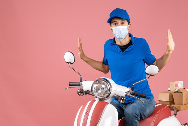 Vista dall'alto dell'uomo corriere confuso in maschera medica che indossa un cappello seduto su uno scooter su sfondo color pesca pastello