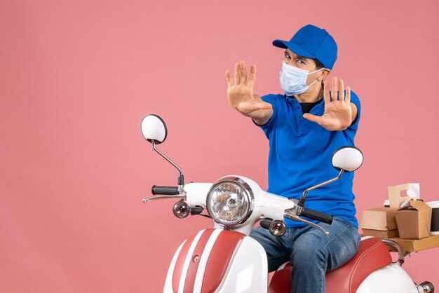 Vista dall'alto dell'uomo corriere in maschera medica che indossa un cappello seduto su uno scooter e mostra dieci su sfondo color pesca pastello