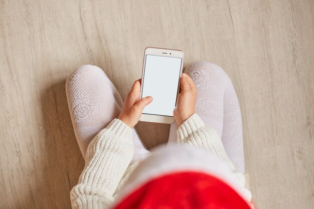 Vista dall'alto della bambina seduta sul pavimento utilizzando uno smartphone, tenendo in mano il cellulare con schermo vuoto per pubblicità o promozione, indossando abiti bianchi e cappello rosso.