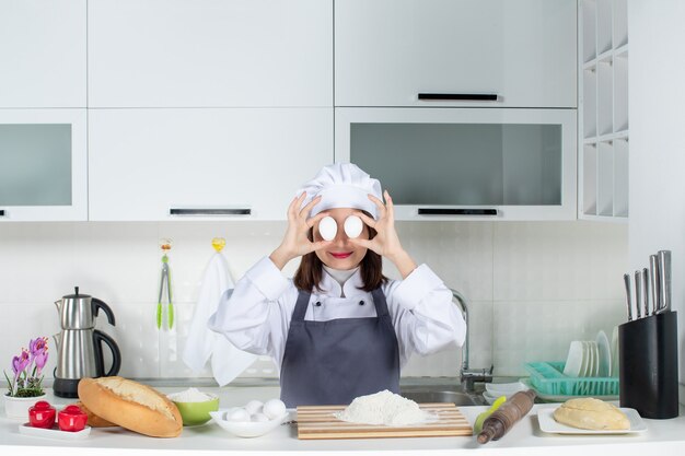 Vista dall'alto della chef donna in uniforme in piedi dietro il tavolo con cibi da tagliere che tengono le uova davanti ai suoi occhi nella cucina bianca