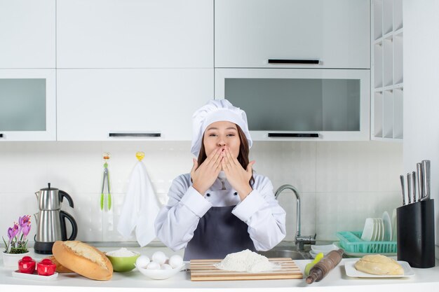 Vista dall'alto della chef donna sorridente in uniforme in piedi dietro il tavolo con le verdure del pane del tagliere che fanno il gesto del bacio kiss